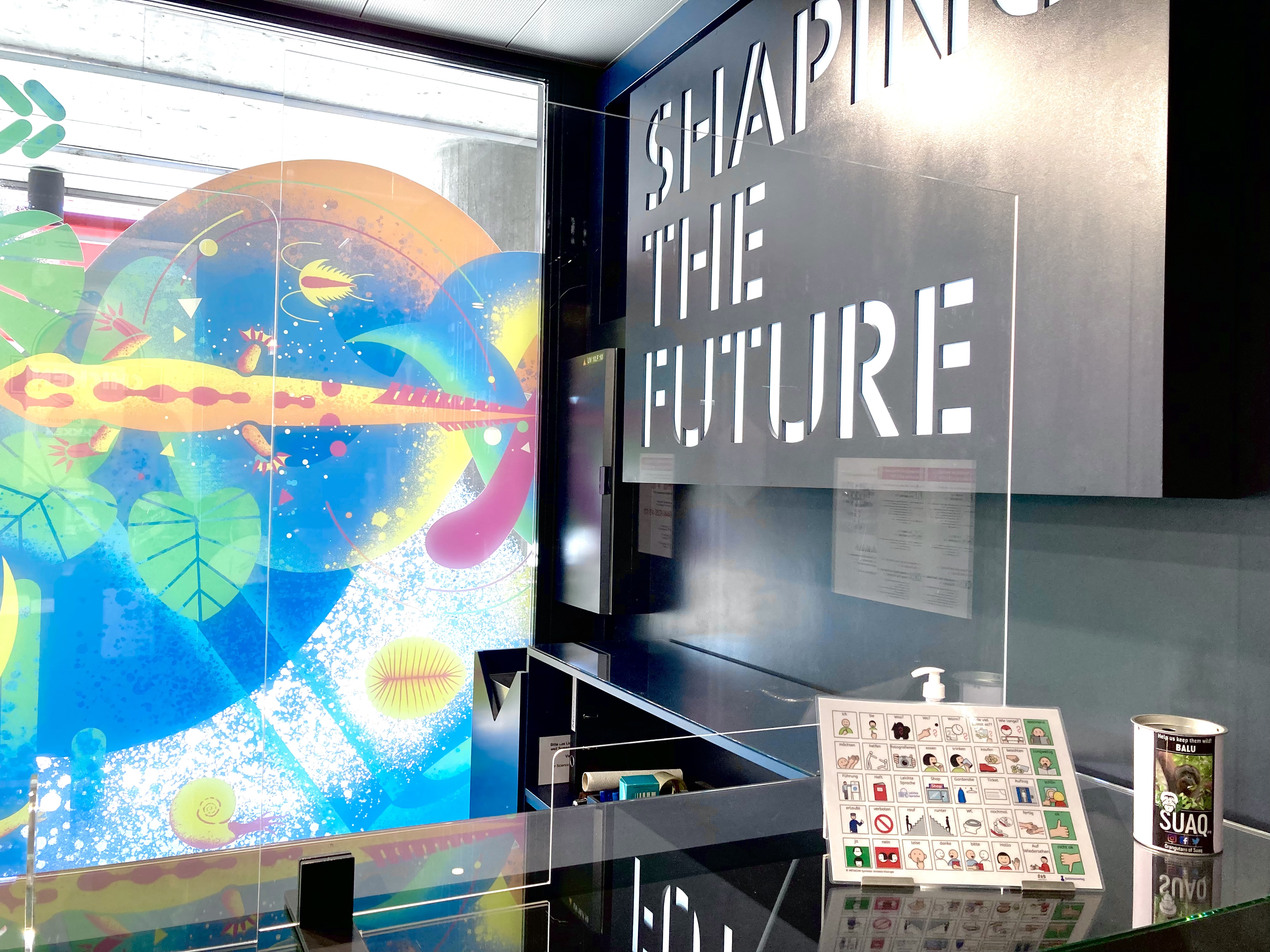 Die Theke des Science Exploratorium UZH mit der Kommunikationstafel. Im Hintergrund ist das bunte Fenster des Museums zu sehen, und am rechten Rand der Schriftzug "Shaping the future".